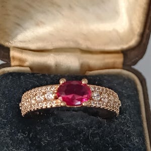 Peut inclure: Une bague en or rose avec une grosse pierre précieuse rouge rubis ovale au centre. La bande est incrustée de petites pierres précieuses claires. La bague est présentée dans un écrin à bijoux vintage.