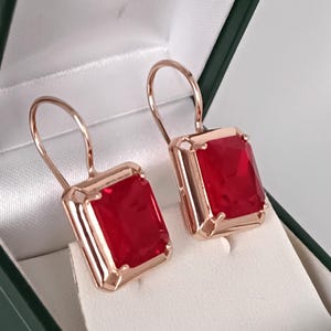 Puede incluir: Un par de pendientes de oro rosa con grandes piedras preciosas rectangulares de color rojo brillante. Los pendientes se muestran dentro de una caja de joyería verde con forro de satén blanco.
