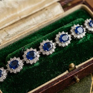 May include: A silver-toned bracelet featuring a series of oval-cut deep blue sapphire gemstones, each surrounded by a halo of small, clear, round-cut white diamonds. The bracelet is arranged in a linear pattern with articulated links.