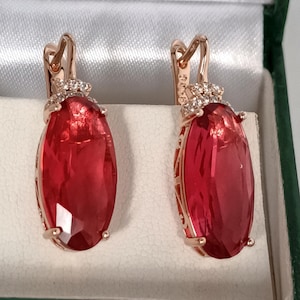 May include: A pair of rose gold earrings with large, oval, red gemstones. The earrings have a lever back closure and are set in a decorative frame with small, clear accent stones. The earrings are displayed in a green and white jewelry box.