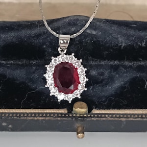 Op de afbeelding: Een zilveren ketting met een hanger met een grote, ovale, dieprode edelsteen omringd door een halo van kleinere, heldere stenen. De hanger hangt aan een fijne zilveren ketting. De ketting wordt gepresenteerd in een zwarte sieradendoos.