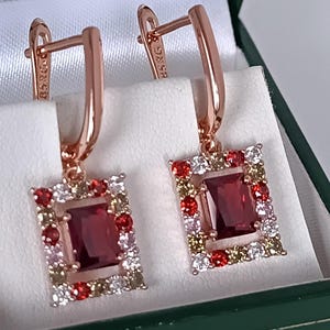 Pode incluir: Brincos em ouro rosa com um centro retangular de pedra preciosa vermelho escuro, rodeado por pedras vermelhas, brancas e amarelas menores. Os brincos são apresentados numa caixa de joias verde.