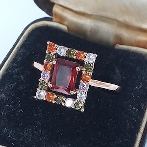 May include: Rose gold ring featuring a square garnet gemstone at the centre, framed by a square of colourful gems in orange, green, white, and pink. The ring is presented in a vintage jewellery box with a black velvet interior.