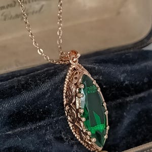 Pode incluir: Um colar em ouro rosa com um grande pendente de pedra verde com corte esmeralda. O pendente está inserido numa moldura decorativa em estilo filigrana. O colar é exibido sobre um fundo escuro e texturizado, realçando os detalhes da joia.