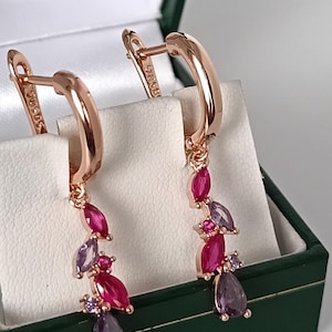 May include: Rose gold earrings with a cascading design. Each earring features pink and purple gemstones. The earrings are presented in a green jewellery box.