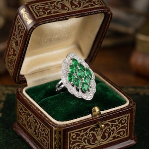 May include: A silver-toned ring featuring a cluster of six marquise-cut green stones arranged in a vertical oval pattern, surrounded by a scalloped border of small clear stones, presented inside a dark brown ornate jewellery box with green velvet.