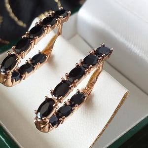 May include: A pair of gold hoop earrings with black oval-shaped gemstones. The earrings are in a jewelry box with a green velvet lining.