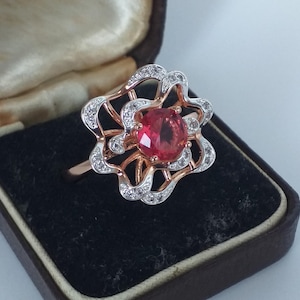 May include: A rose gold ring with a large, round, red gemstone at the center, surrounded by a floral design with white gemstones. The ring is displayed in a vintage jewelry box.