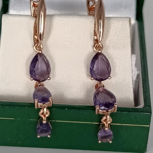 Peut inclure: Une paire de boucles d'oreilles pendantes en or rose composées de trois pierres précieuses violettes facettées en forme de poire, disposées verticalement par taille décroissante et suspendues à une attache dormeuse en or rose.