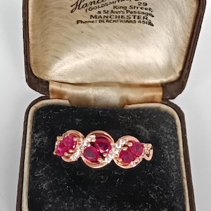 Op de afbeelding: Een roségouden ring met drie ovale robijnen en kleine witte edelstenen. De ring wordt gepresenteerd in een vintage bruine sieradendoos met de tekst "Hancock & Jon" en "Manchester".