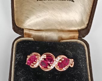 May include: A rose gold ring with three oval-cut rubies and small white gemstones. The ring is displayed in a vintage brown jewelry box with the text "Hancock & Jon" and "Manchester" visible.
