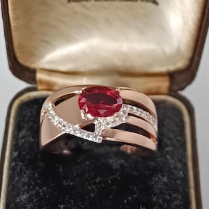May include: Rose gold ring featuring a large oval ruby center stone. The ring has a unique design with diamond accents and a split shank. The ring is displayed in a vintage jewelry box.