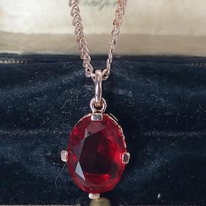 May include: A rose gold necklace featuring a large, oval-cut, red gemstone pendant. The pendant is set in a rose gold frame and suspended from a fine chain. The necklace is presented in a vintage jewellery box.