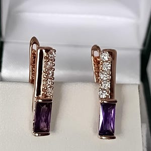 Peut inclure: Une paire de boucles d'oreilles couleur or rose avec une pierre précieuse rectangulaire violette à la base et une rangée de quatre petites pierres rondes transparentes sur la partie supérieure, dotées d'un fermoir à levier articulé.