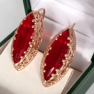 May include: A pair of gold-toned earrings with large, oval, red gemstones. The earrings have an ornate, filigree design around the gemstones and are displayed in a white-lined box with a green exterior.
