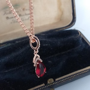 May include: Rose gold necklace with a teardrop-shaped, deep red gemstone pendant. The pendant is accented with small, clear crystals. The necklace is displayed in a vintage black jewelry box.