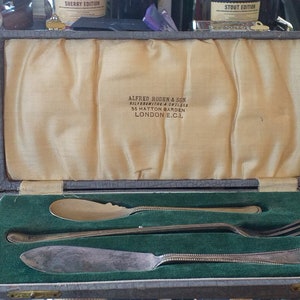 May include: A silver cutlery set in a brown velvet-lined box. The box is open and the cutlery is visible. The cutlery includes a butter knife, a spoon, and a fork. The box is labelled "Alfred Baker & Son Silversmiths & Jewellers 35 Hatton Garden London E.C.1."