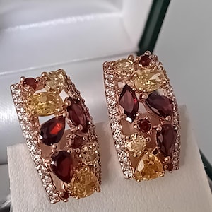 May include: A pair of rectangular rose gold earrings featuring a cluster of faceted pear and oval-shaped deep red garnet and yellow citrine gemstones, accented by small clear white pavé stones along the outer edges of the metal frame.