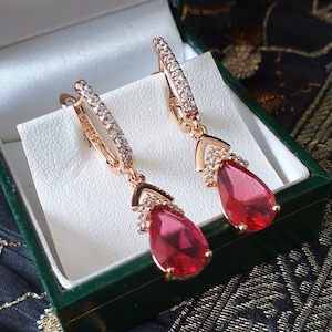 May include: A pair of rose gold earrings with teardrop-shaped red gemstones and clear cubic zirconia accents. The earrings are displayed in a green velvet jewelry box.