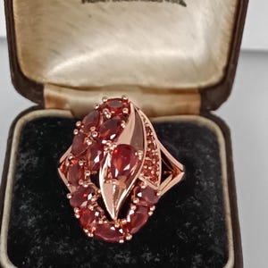 May include: A rose gold ring with a cluster of oval and pear-shaped red gemstones. The ring is set in a decorative design and is displayed in a brown jewelry box with a black velvet interior.