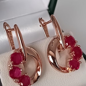 May include: A pair of rose gold hoop earrings featuring a circular design accented with three round, deep red rubies on each piece. The earrings have a hinged lever back closure and are stamped with the text 585RG on the metal surface.