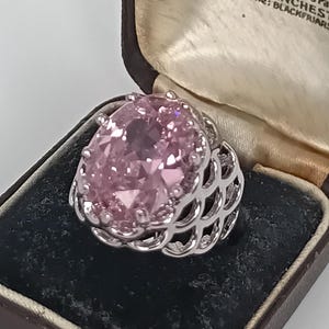 Könnte beinhalten: Ein silberfarbener Ring mit einem großen, oval geschliffenen, hellrosa Edelstein in einer Krappenfassung. Die Ringschiene weist ein kompliziertes, durchbrochenes, bogenförmiges Metalldesign auf. Der Ring liegt in einer dunkelbraunen Schmuckschatulle.