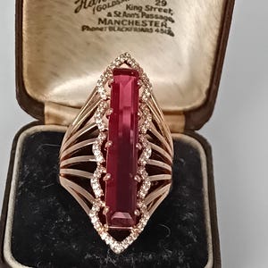 May include: A rose gold ring featuring a long, rectangular, deep red gemstone set vertically. The stone is framed by a scalloped border of small, clear, sparkling stones. The band consists of multiple thin, parallel rose gold wires on each side.
