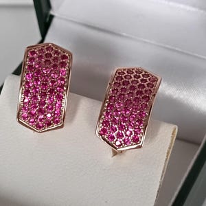 Puede incluir: Un par de pendientes de oro rosa con forma de escudo. Los pendientes están incrustados con numerosas piedras preciosas pequeñas de color rosa brillante. Se presentan en una caja de joyería blanca con exterior verde.