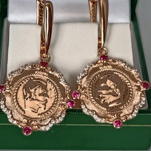 Könnte beinhalten: Ein Paar Ohrhänger aus Roségold mit runden münzartigen Anhängern mit geprägten Profilen. Jeder Anhänger ist von einem gewellten Rand mit kleinen klaren Steinen umgeben und mit vier runden, leuchtend pinkfarbenen Edelsteinen an den Seiten akzentuiert.
