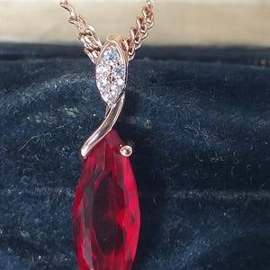 Könnte beinhalten: Halskette aus Roségold mit einem großen, facettierten, tropfenförmigen roten Edelsteinanhänger. Der Anhänger ist mit einer kleineren, oval geformten Ansammlung klarer Steine versehen. Die Kette ist filigran.