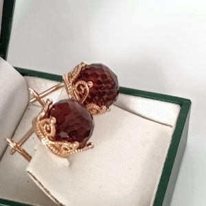 May include: A pair of gold-toned cufflinks with faceted, dark red gemstone spheres. The cufflinks are displayed in a green box with a white interior, showcasing their intricate design and craftsmanship.