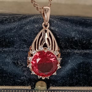 Könnte beinhalten: Halskette mit roségoldfarbenem Anhänger, der einen großen, runden, roten Edelstein trägt. Der Edelstein ist von kleinen, klaren Steinen umgeben und in einem verzierten, durchbrochenen, tropfenförmigen Rahmen gefasst. Die zarte Kette passt zur Farbe des Anhängers.