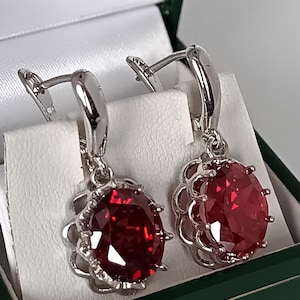 May include: A pair of silver-toned drop earrings featuring oval-cut, deep red faceted stones. Each stone is secured in a decorative, scalloped metal setting with a hinged lever-back closure, presented in a dark green jewelry box.