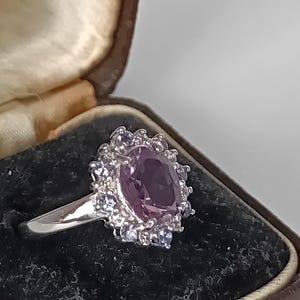 May include: A silver-toned ring featuring a large, oval-cut purple gemstone at the center, surrounded by a halo of small, clear, round-cut stones. The ring has a polished metal band and is presented in a dark-lined, hinged jewelry box.