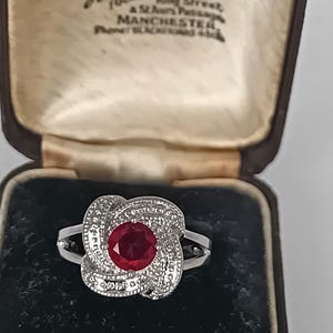 Può includere: Un anello color argento con una pietra preziosa rossa dal taglio rotondo al centro, circondata da un design in metallo testurizzato a vortice. L'anello è presentato in un portagioie marrone scuro con interno color crema e scritta Hancock & Son.