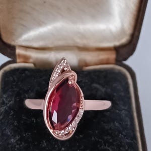 May include: Rose gold ring with a large, marquise-cut, deep red gemstone. The ring features a delicate design with small, clear accent stones. Presented in a vintage jewelry box with a satin lining.