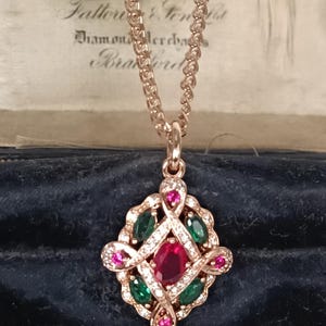 Puede incluir: Collar con colgante de oro rosa con un rubí central ovalado, rodeado de esmeraldas, gemas rosas y pequeños diamantes. El colgante cuelga de una delicada cadena. El fondo es de terciopelo azul oscuro.