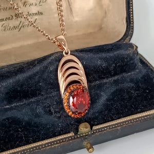 Op de afbeelding: Rosegouden hangerketting met een ovale, rode edelsteen omringd door kleinere oranje stenen. De hanger heeft een gelaagd ontwerp en een fijne ketting. De ketting wordt gepresenteerd in een vintage sieradendoosje met een donkerblauwe binnenkant.