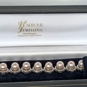 May include: A bracelet featuring a series of round, light pink pearls, each set within a circular silver-toned frame accented with small clear stones. The jewellery is presented in a black display box with the text Harbour Jewellers Mullingar printed inside.