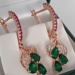 Könnte beinhalten: Ohrringe aus Roségold mit Blattdesign. Die Reifen sind mit kleinen roten Steinen besetzt. Jeder Ohrring hat drei grüne tropfenförmige Steine und kleine klare Steine.