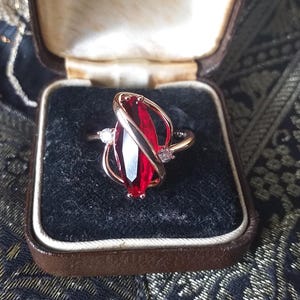 Könnte beinhalten: Ein Ring mit einem roten Edelstein, einem goldenen Band und zwei kleineren Diamanten an den Seiten. Der Ring befindet sich in einer samtenen Schachtel mit der Aufschrift "Hancock & Son (Goldsmiths) 29 King Street, St Ann's Passage, Manchester, Phone: BLACKFRIARS 456".