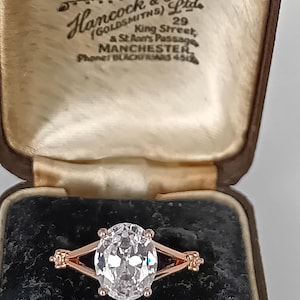 May include: An oval-cut clear gemstone ring set in a rose gold band with a split shank design, presented in a brown velvet-lined jewellery box with the text Hancock & Son Ltd, Diamond Merchants, 29 King Street, St Ann's Passage, Manchester printed inside.