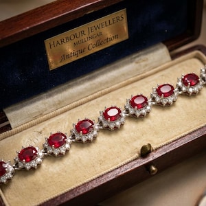 May include: A bracelet featuring oval-cut red gemstones surrounded by small white stones, presented in a dark wooden box with a gold plaque reading HARBOUR JEWELLERS MULLINGAR Antique Collection. The bracelet consists of multiple linked segments.