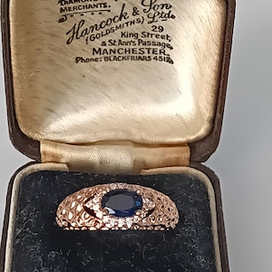 May include: A rose gold ring featuring a dark blue oval gemstone and small white stones, set in an intricate filigree pattern. The ring is presented in a vintage brown jewellery box. The box has text that reads "Hancock & Son Ltd, Diamond Merchants, Manchester".
