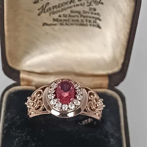 Puede incluir: Un anillo de oro rosa con una piedra preciosa roja de corte ovalado central rodeada por un halo de pequeños diamantes redondos. La banda tiene un intrincado trabajo de filigrana en los lados. El anillo se presenta en un joyero.