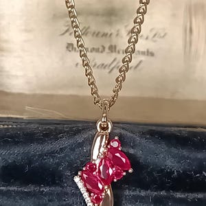 Könnte beinhalten: Goldfarbene Halskette mit Anhänger. Der Anhänger hat einen Cluster aus oval geschliffenen, tiefroten Edelsteinen, kleinen, klaren Edelsteinen und einem geschwungenen Golddesign. Die Halskette wird vor einem dunkelblauen Hintergrund präsentiert.