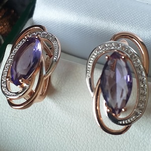 Op de afbeelding: Een paar oorbellen van roségoud met een paarse amethist edelsteen die in elke oorbel is gezet. De oorbellen zijn versierd met een halo van kleine witte diamanten.