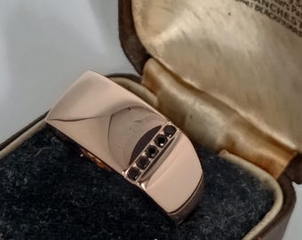 Könnte beinhalten: Ein roségoldener Ring mit rechteckigem Design und einer Reihe kleiner, dunkler Edelsteine. Der Ring wird in einer Vintage-Schmuckschatulle mit Samtinnenraum und gedrucktem Etikett präsentiert.