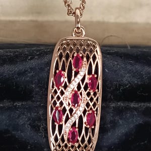 Op de afbeelding: Rosegouden hangerketting met een rechthoekig openwerkontwerp. De hanger is versierd met ovaal geslepen rode edelstenen en een sprankelend wit edelsteenaccent. De ketting wordt gepresenteerd in een donkerblauwe fluwelen sieradendoos.