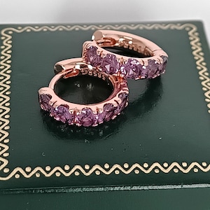 Op de afbeelding: Een paar roségouden oorringen, elk bezet met kleine, ronde, roze edelstenen. De oorbellen worden gepresenteerd op een donkergroene doos met een gouden decoratieve rand. Een klassiek sieraad.